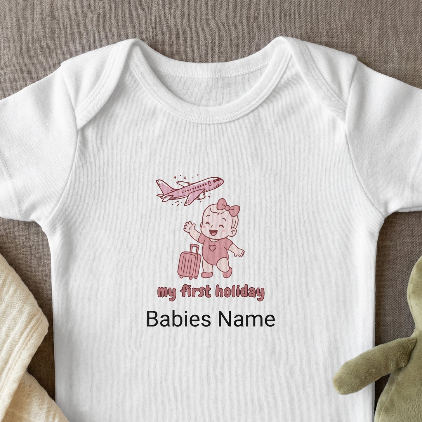White baby onesie with pink "my first holiday" text, cartoon baby, and airplane.