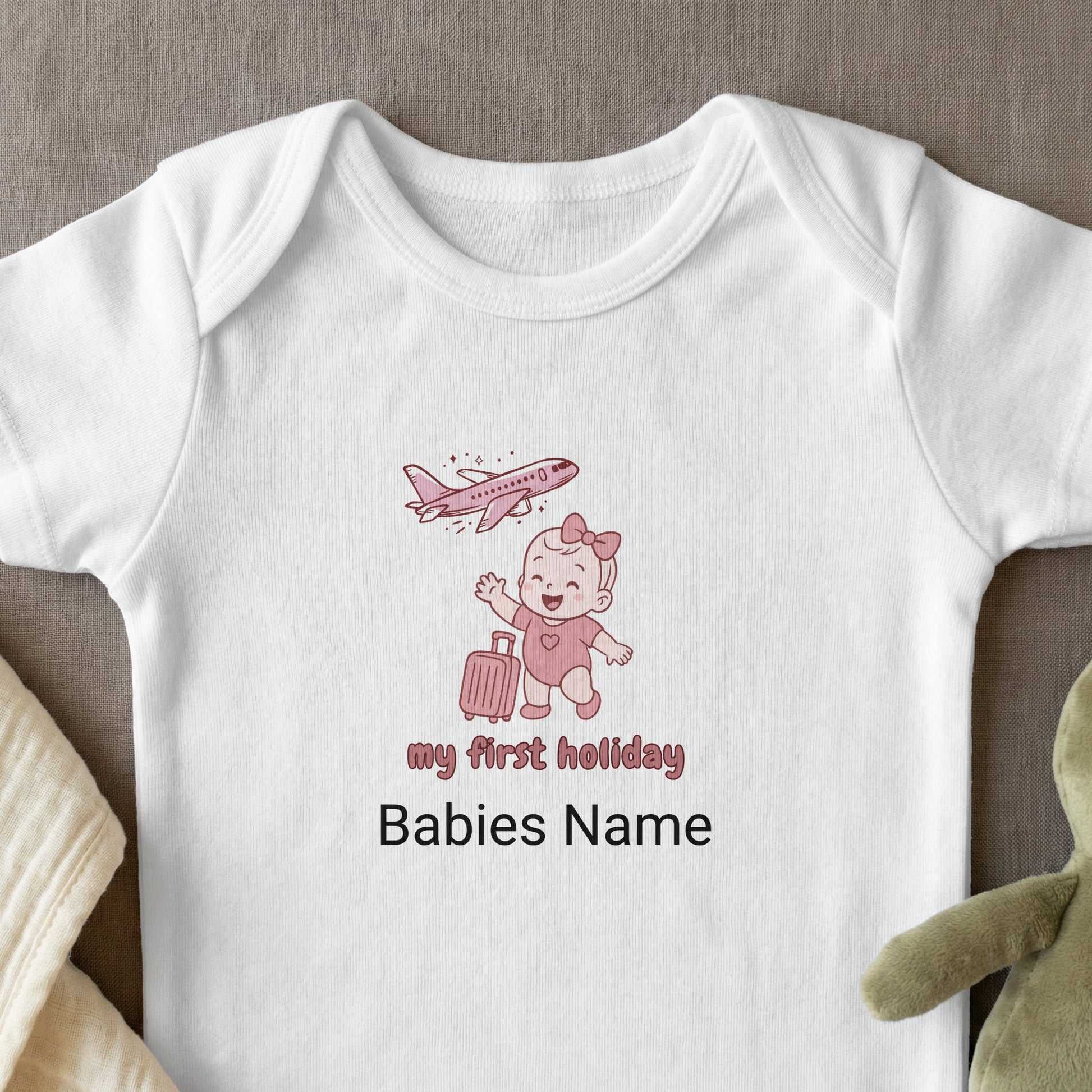 White baby onesie with pink "my first holiday" text, cartoon baby, and airplane.