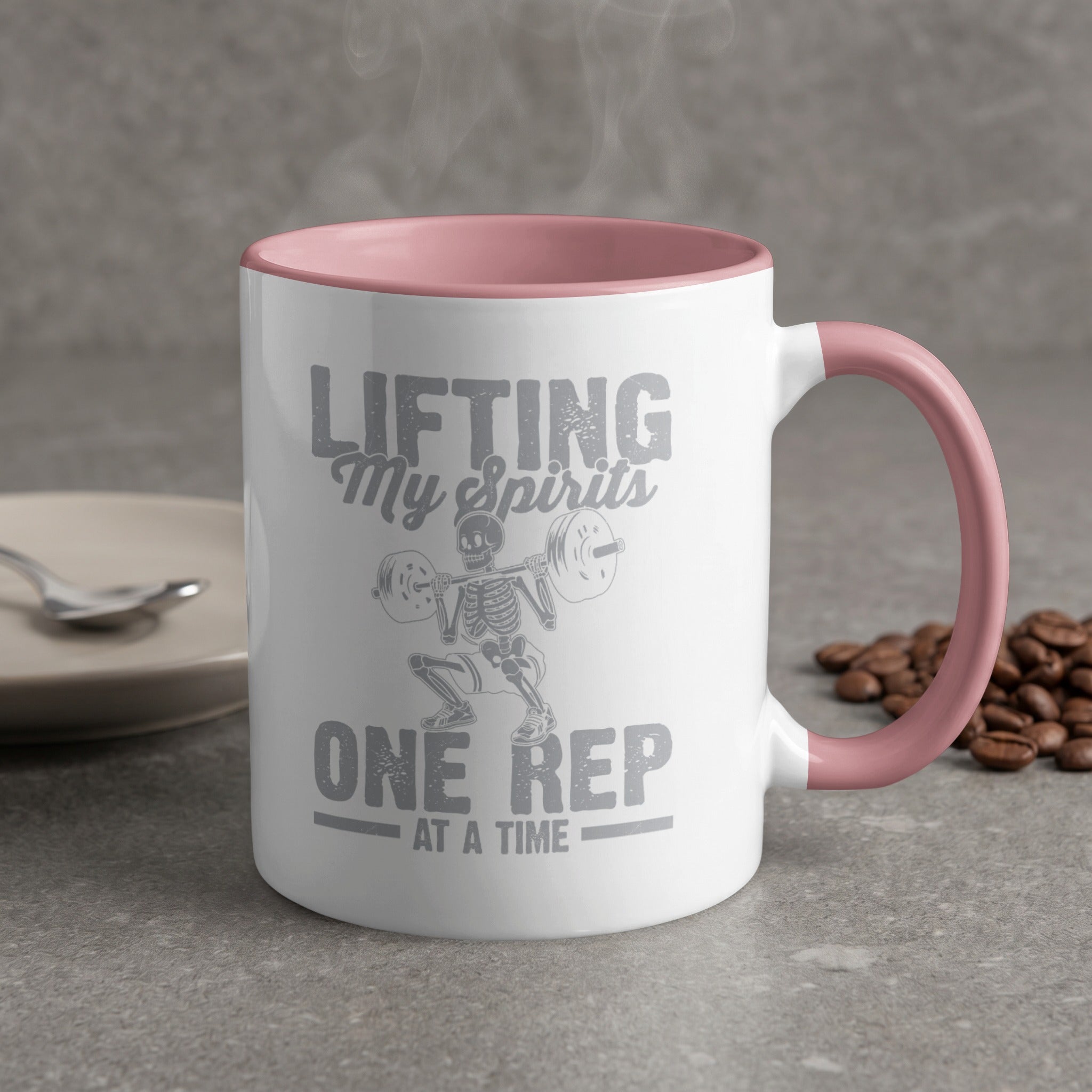 White mug with pink handle, featuring a skeleton lifting weights and the text "Lifting My Spirits ONE REP AT A TIME".