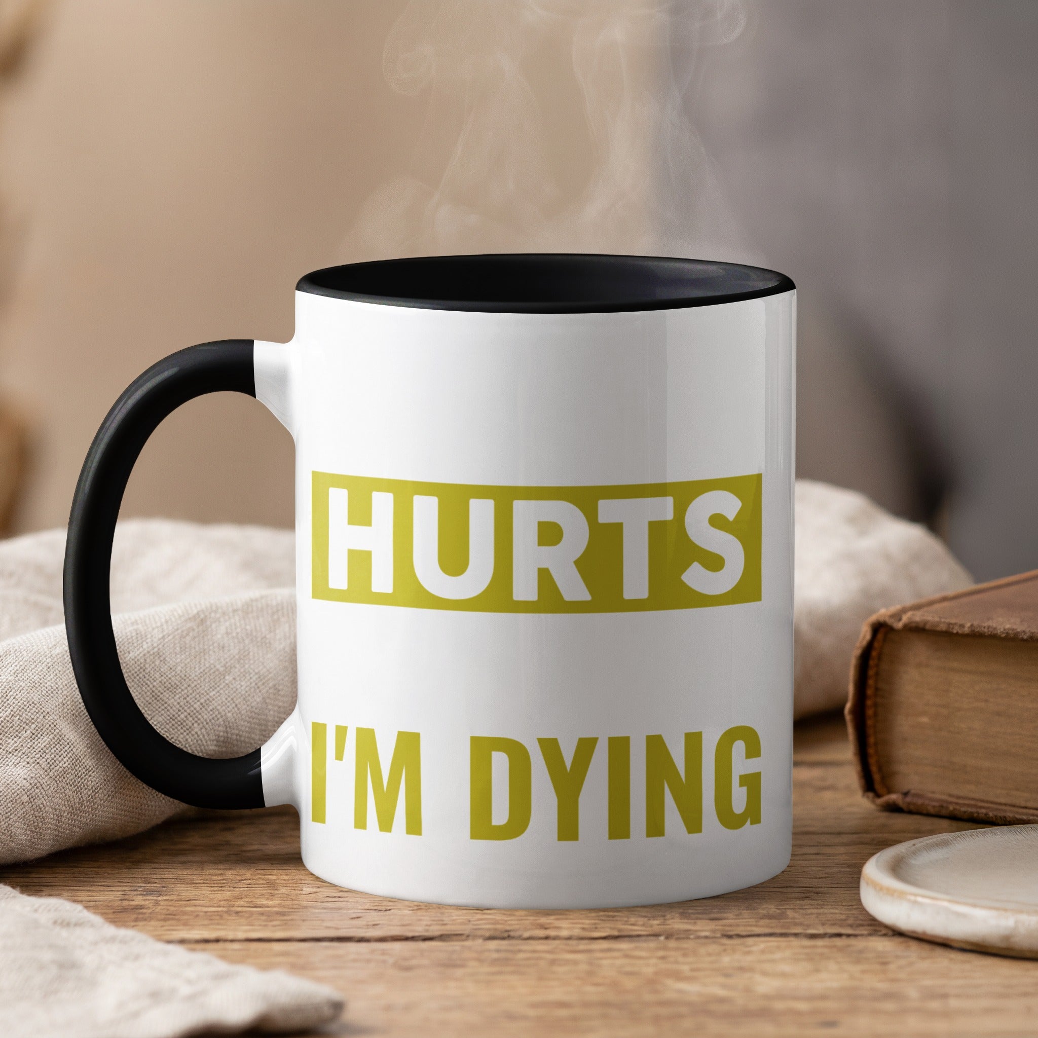 White mug with black handle, steaming, displaying text "HURTS I'M DYING".