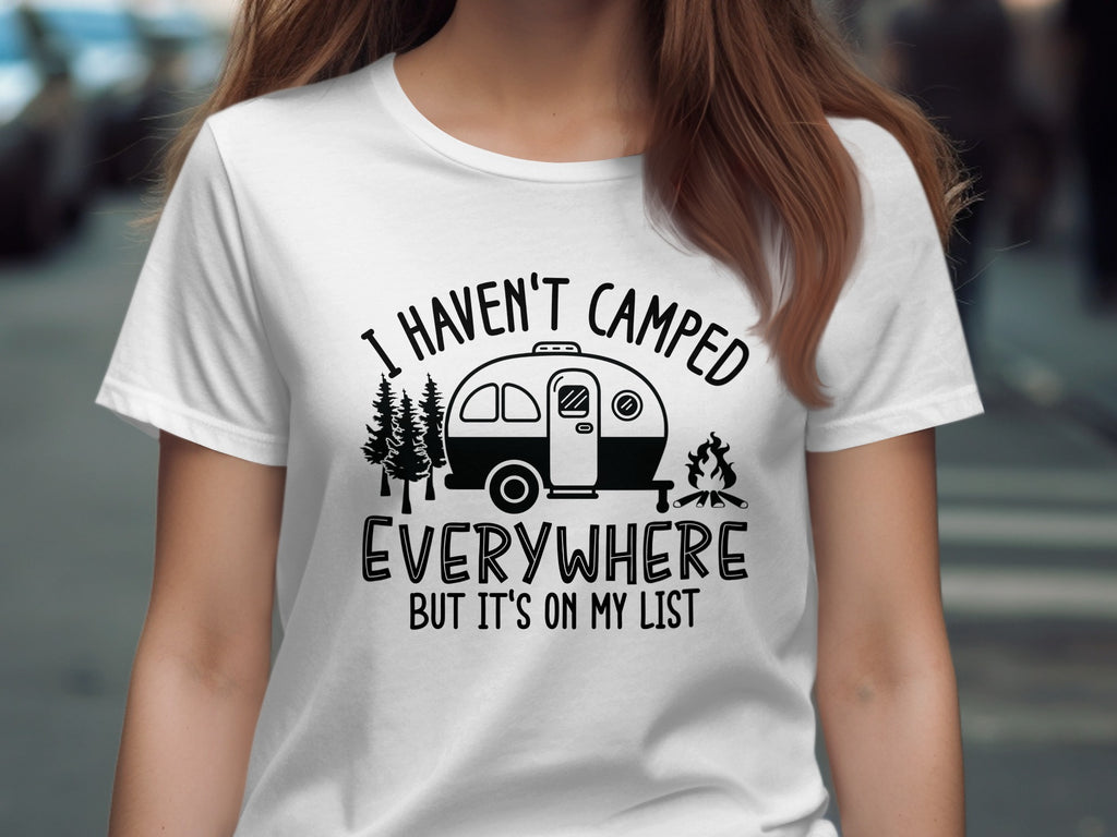 White t-shirt with black graphic featuring a camper, trees, campfire, and text "I HAVEN'T CAMPED EVERYWHERE BUT IT'S ON MY LIST".